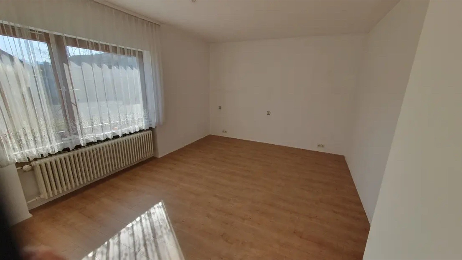 Leeres Wohnzimmer mit hellem Holzfußboden, Heizkörper und großen Vorhängen